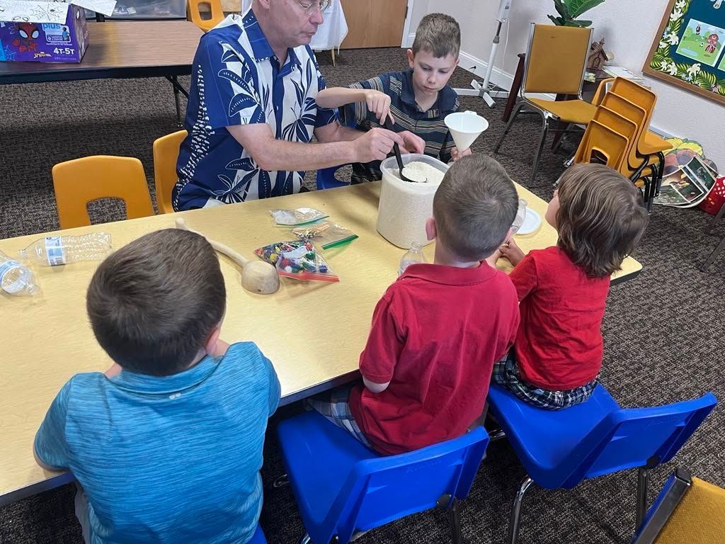 Adult serving food to three children seated at a table in a classroom or daycare.