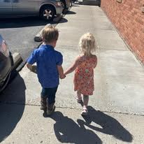 Two children holding hands while walking on a sunny sidewalk beside parked cars and a brick wall