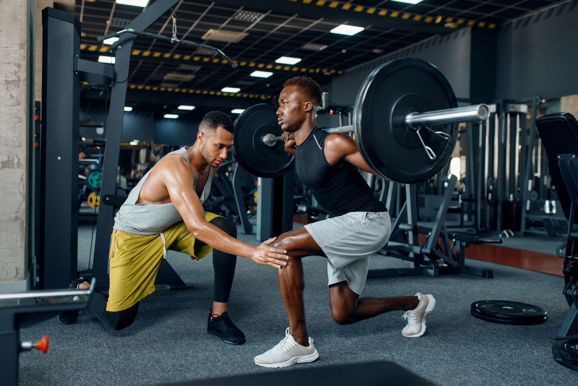 Two men training with a barbell in a gym, one spotting the other during a lunge.