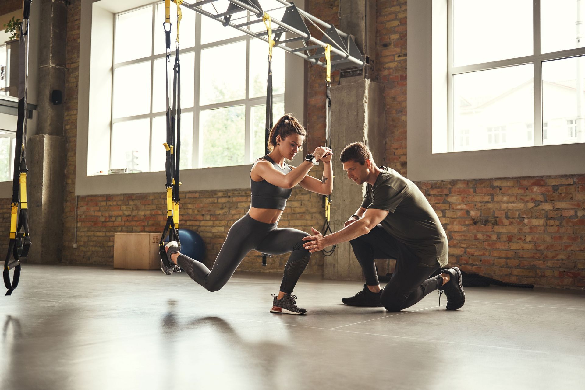 Two people exercising in a gym, one kneeling and assisting a woman on suspension straps in front of a brick wall.