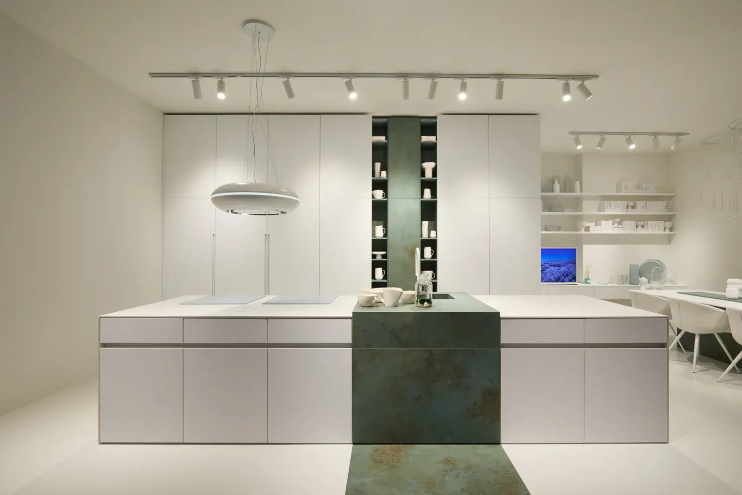 Moderna cucina bianca con ampia isola, dettagli color verde acqua e illuminazione dall'alto. Sullo sfondo si intravedono un piccolo spazio di lavoro e degli scaffali.