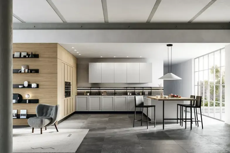 Cucina moderna con mobili in legno chiaro, ripiani bianchi e pavimento in pietra. Include un'isola con sgabelli da bar e una morbida poltrona grigia.