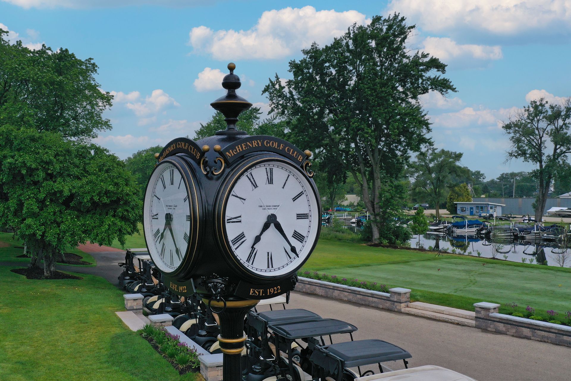 A vintage-style, double-faced black clock stands on a post overlooking a golf course with green grass and trees.