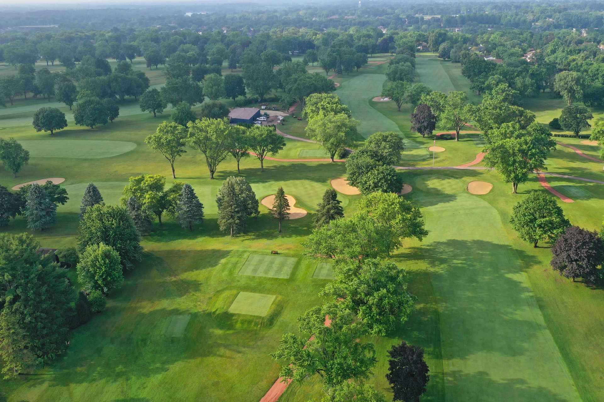 An aerial view of a lush green golf course with fairways, sand traps, and surrounding trees on a sunny day.