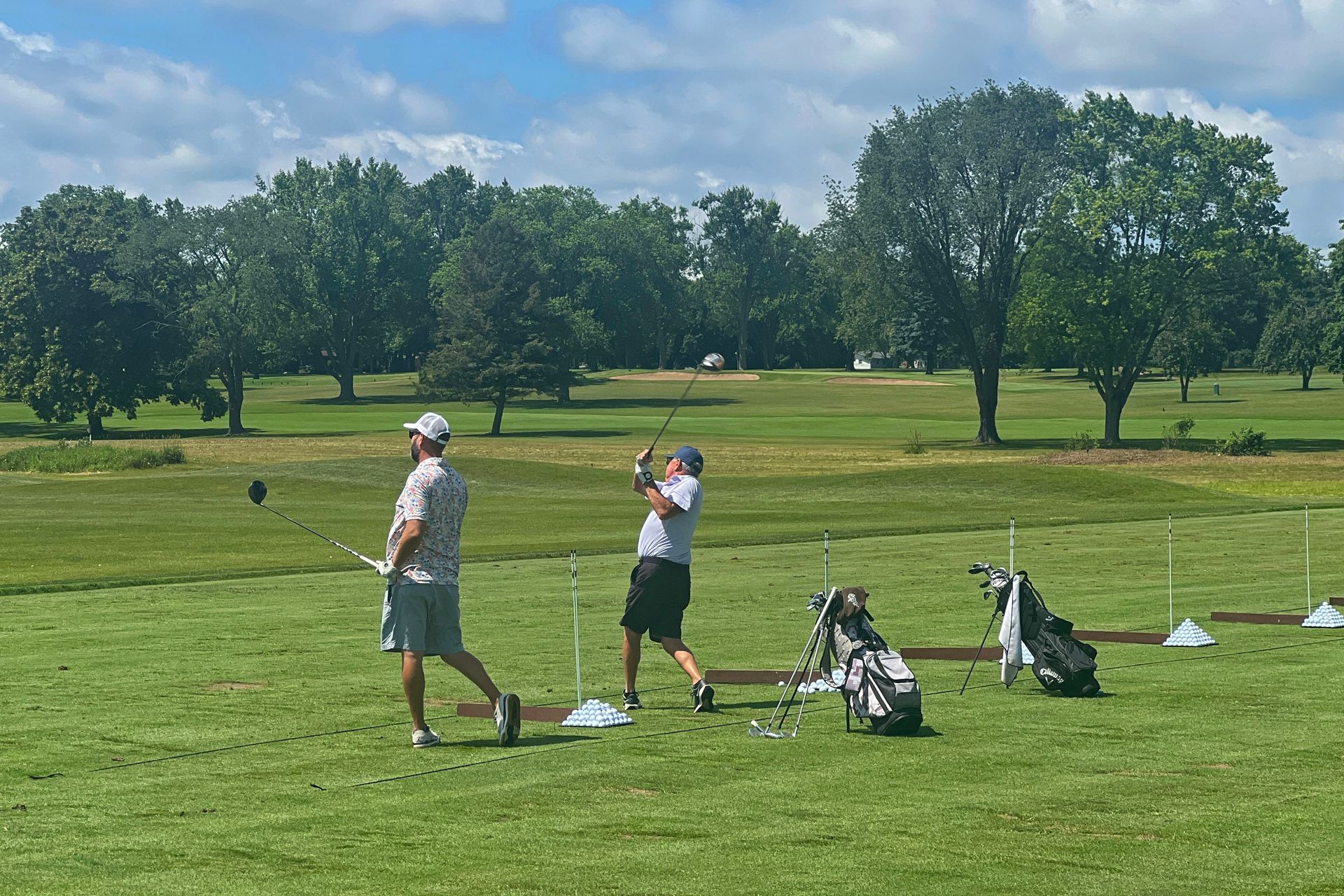 Two people stand on a lush green golf course driving range, one mid-swing with a club and another observing.