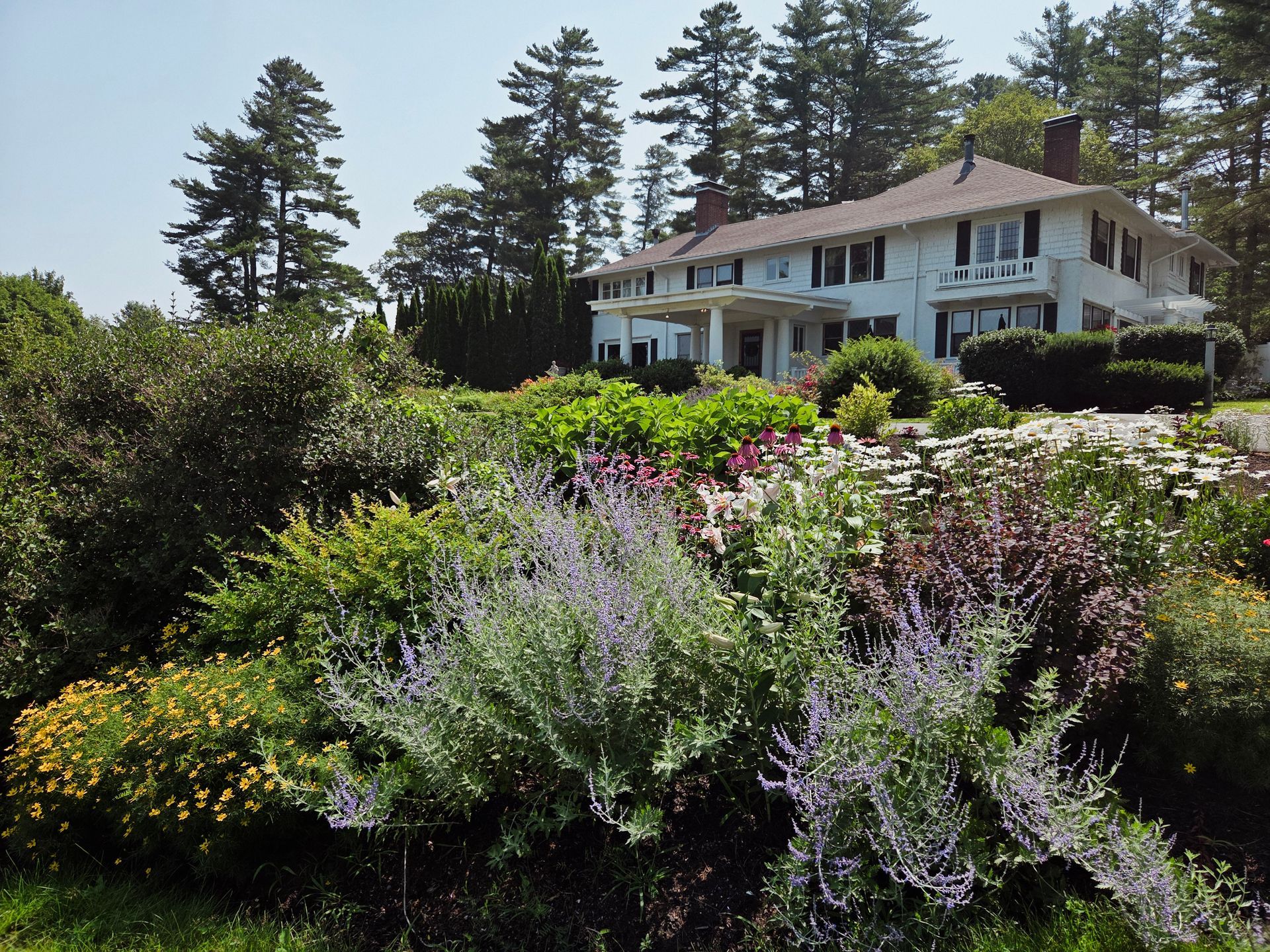 Large white house behind lush flower garden and tall trees on a sunny day