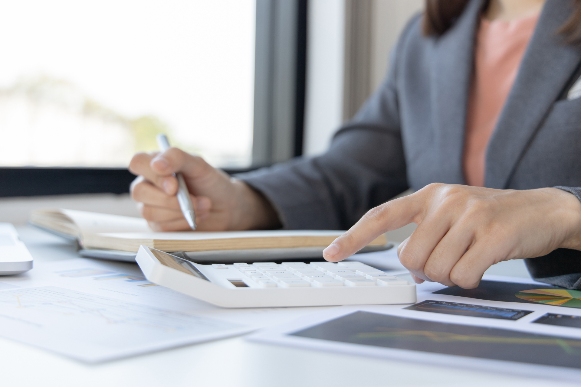 Person in gray blazer using a calculator and taking notes at a desk near a window.