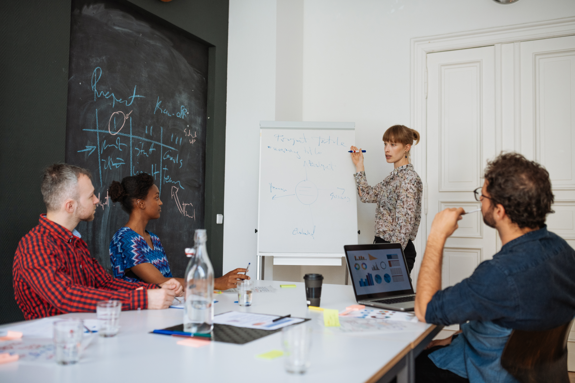 Four people in meeting. Woman writes on whiteboard, while others look on, seated around a table.