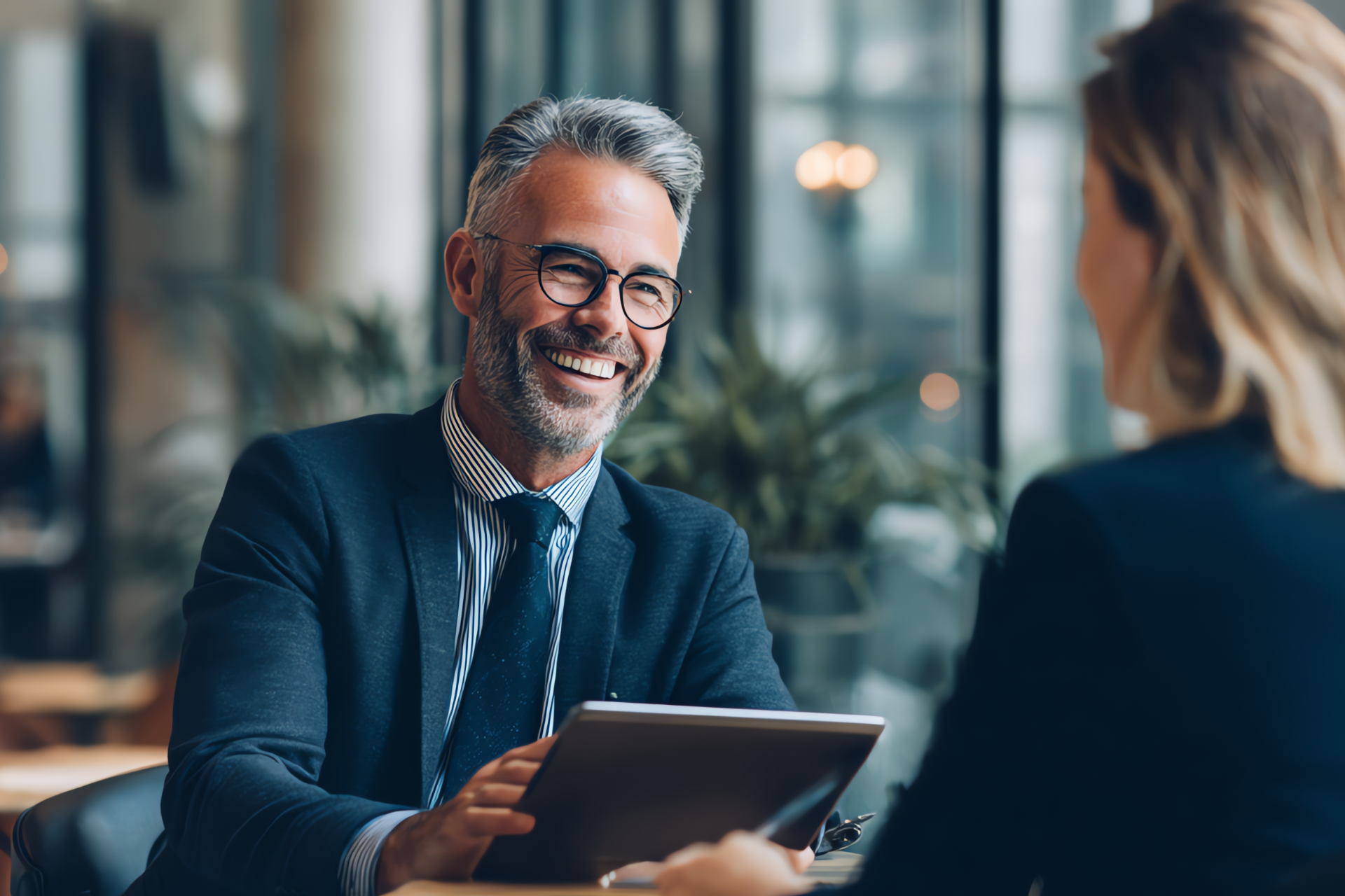 Man in suit smiles at person while holding tablet in an office setting.