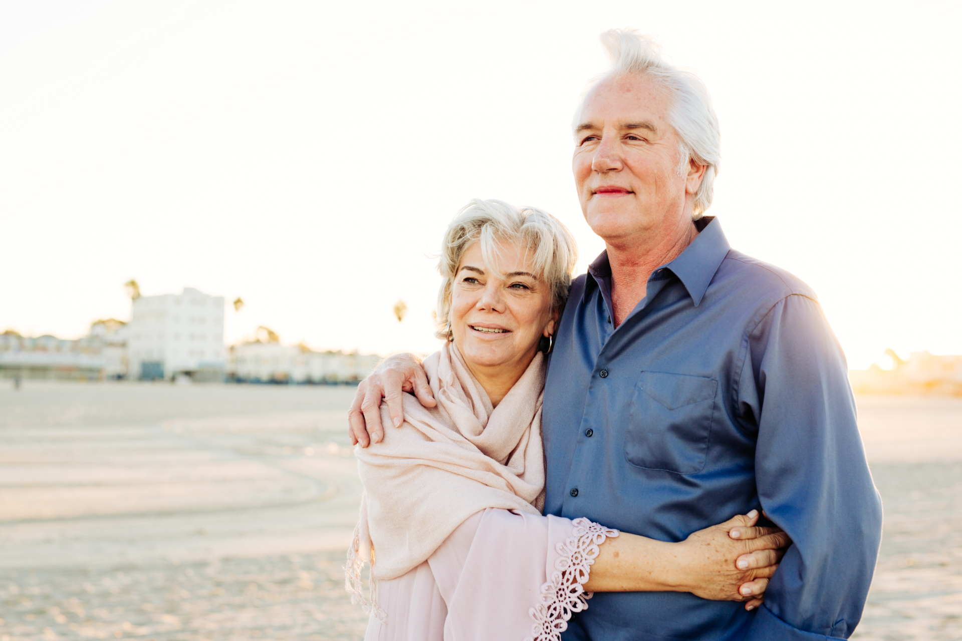Elderly couple embracing on a beach, looking towards the sun.