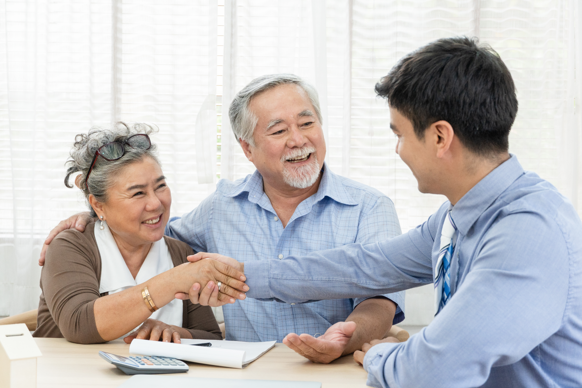 An older couple shakes hands with a younger person at a table, smiling. Bright indoor setting.