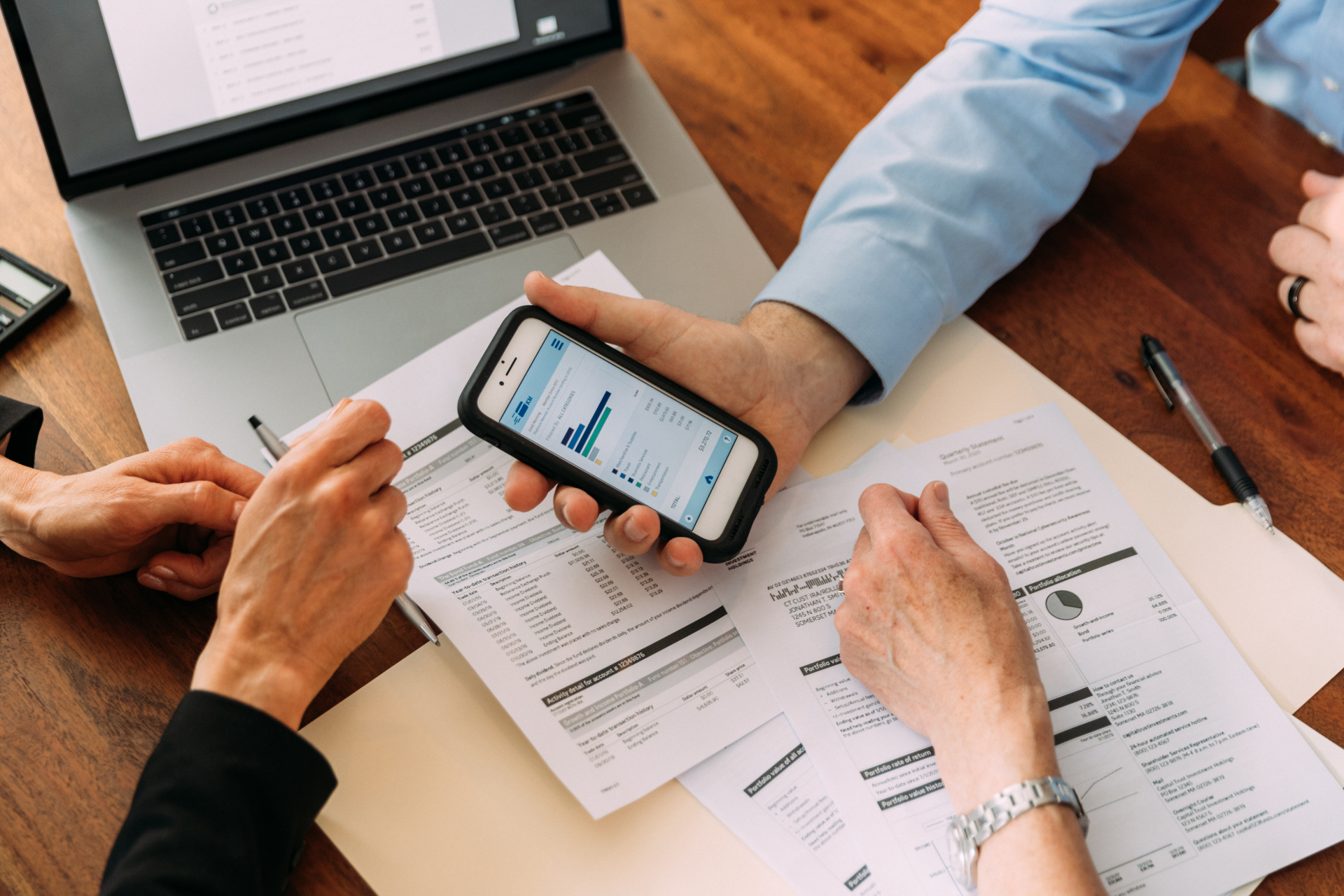 Hands examining financial documents, smartphone with chart, laptop on desk.