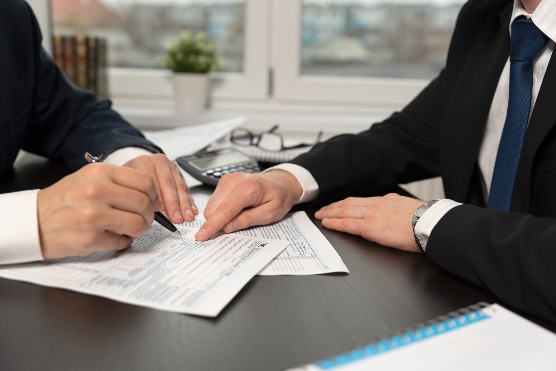 Two men in suits reviewing paperwork, one signing, near a calculator and window.