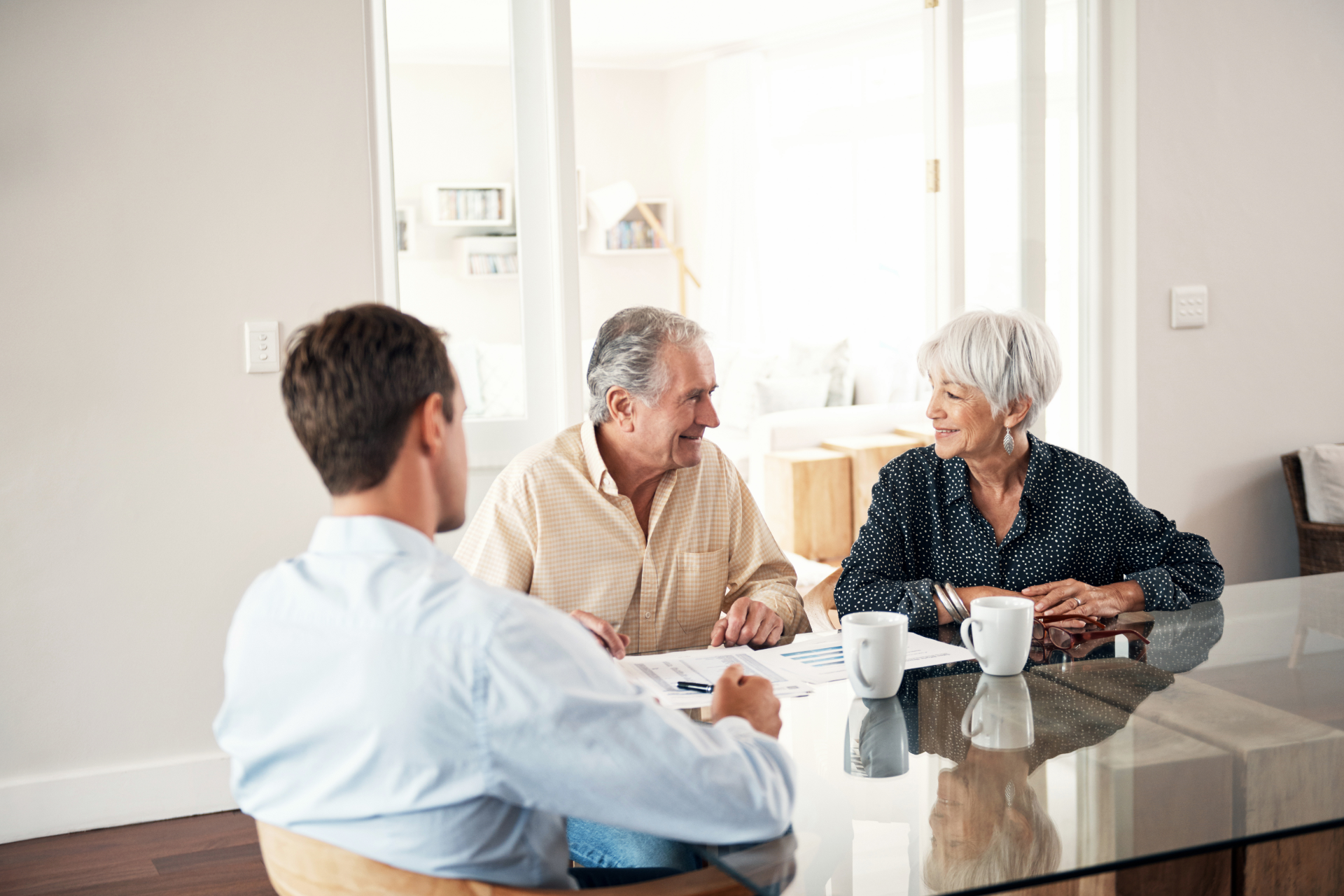 A man in a light blue shirt advises an older couple, seated at a table with papers, inside a home.
