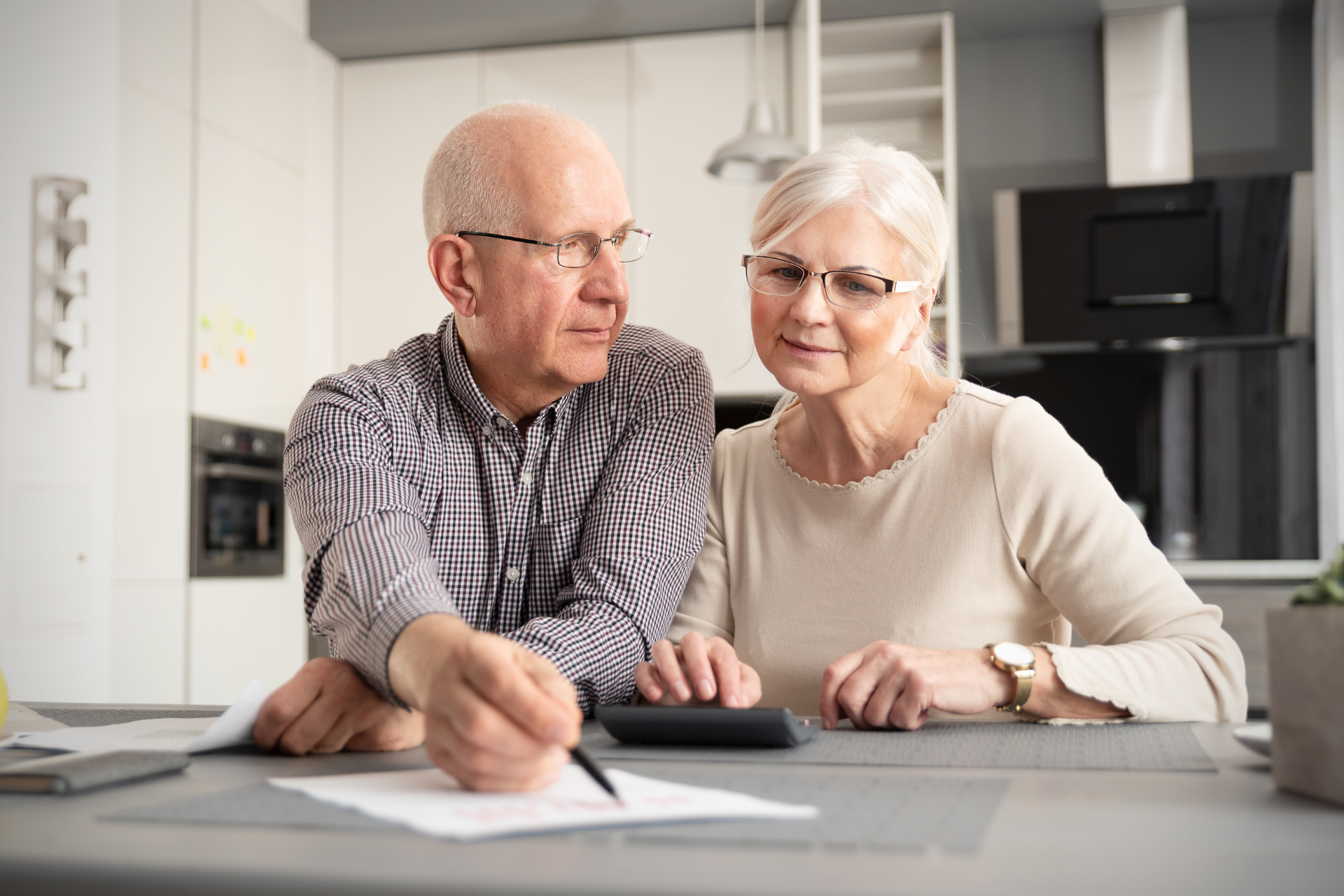 Elderly couple reviewing documents and using a calculator in a kitchen.
