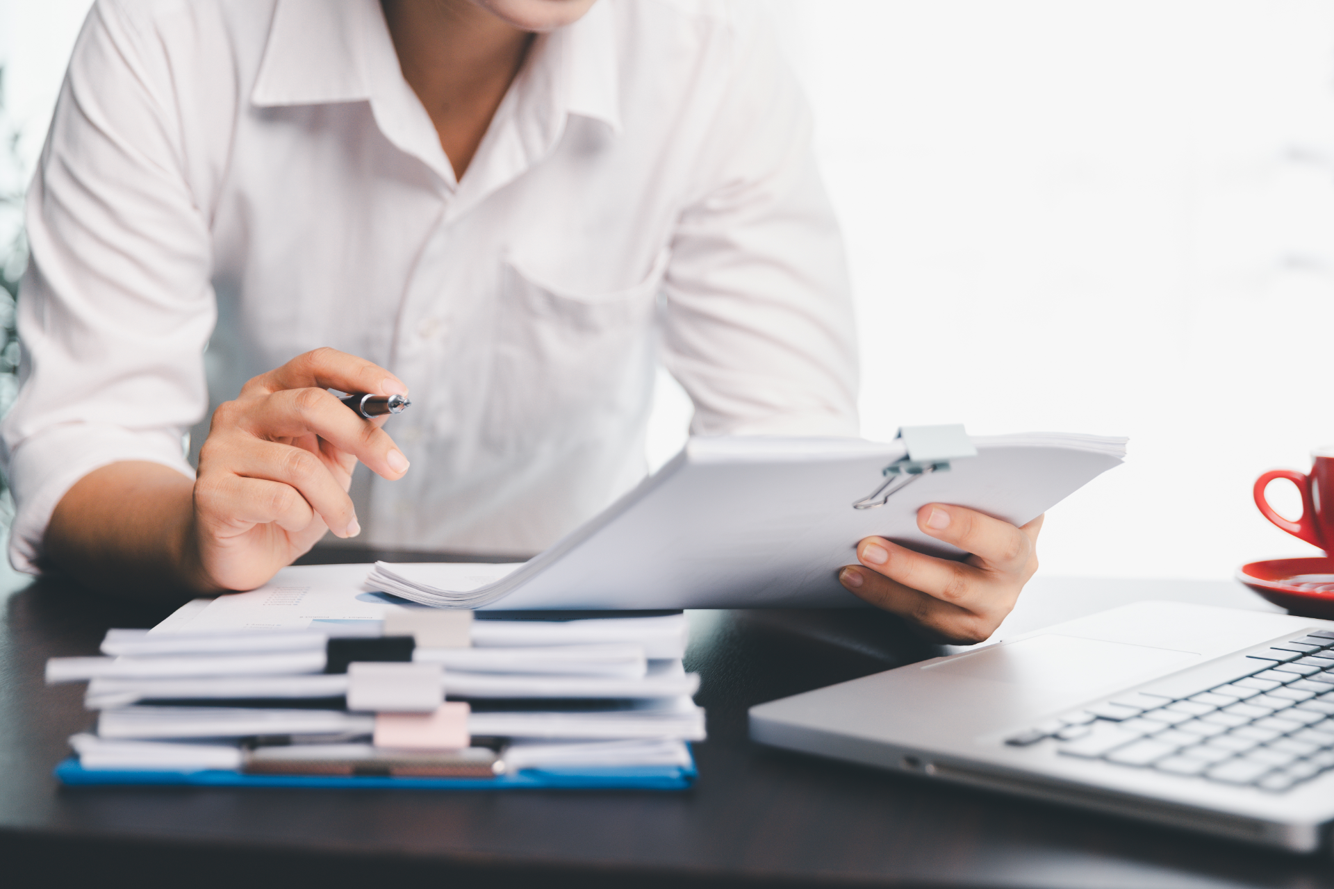 Person in white shirt reviewing documents at a desk, laptop and coffee nearby.
