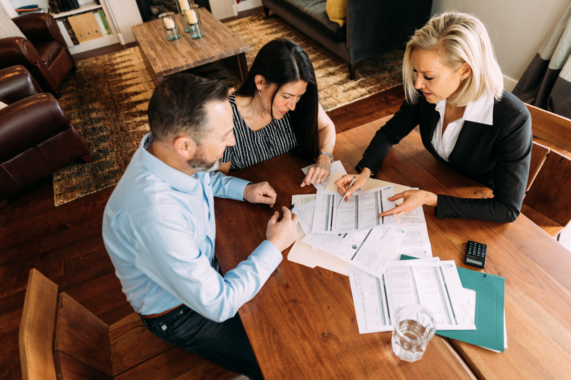 Couple and financial advisor reviewing paperwork at a wooden table. Blonde woman pointing, looking at documents.