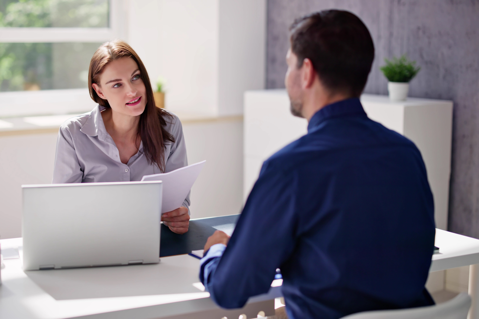 Woman interviewing a person at a desk; she holds papers, laptop, and is speaking.