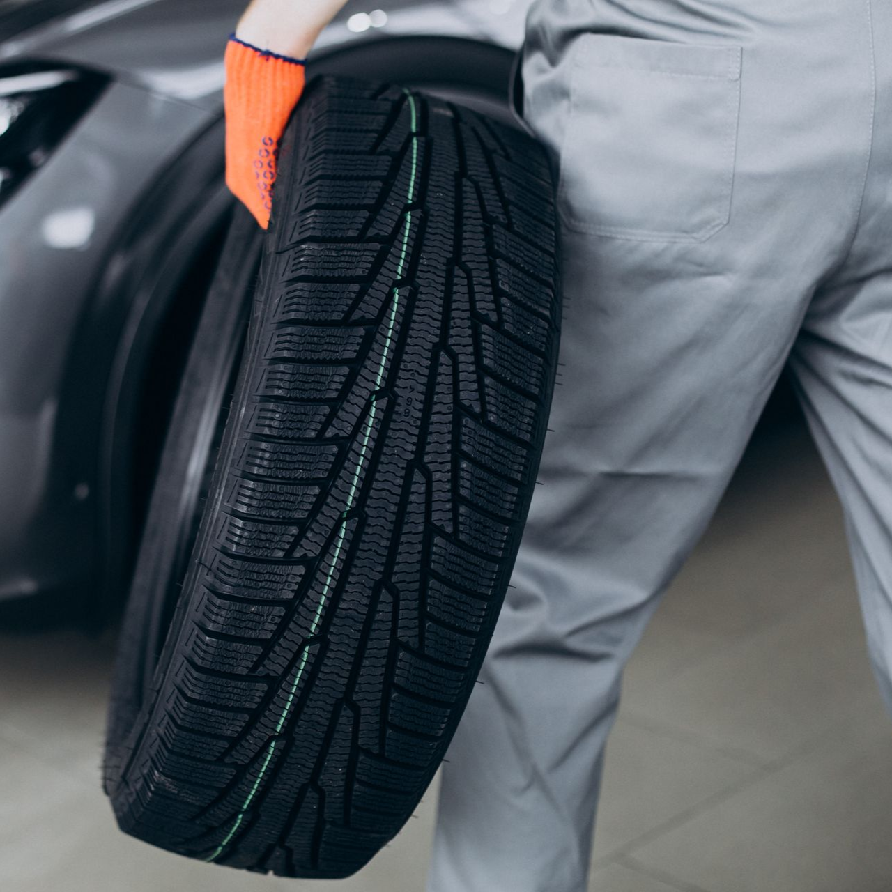 Mechanic in gray overalls holding a tire, orange glove visible, next to a car.