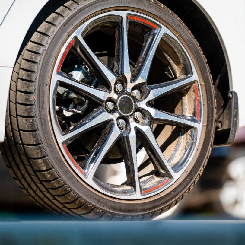 Close-up of a car wheel with a black and silver rim, mounted on a white car, resting on asphalt.