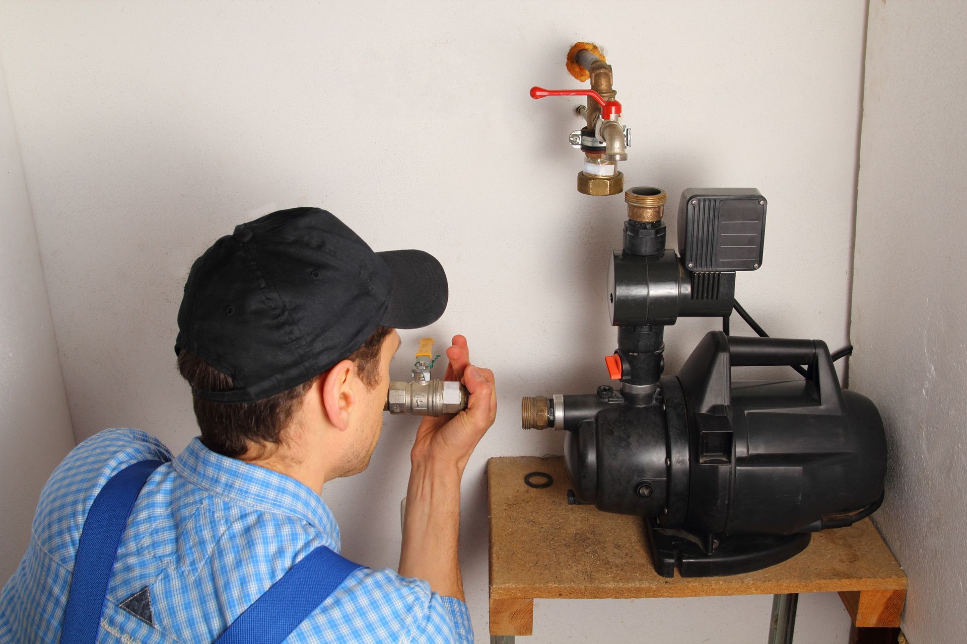 A technician in workwear inspecting a plumbing valve near a black water pump installed on a wooden stand.