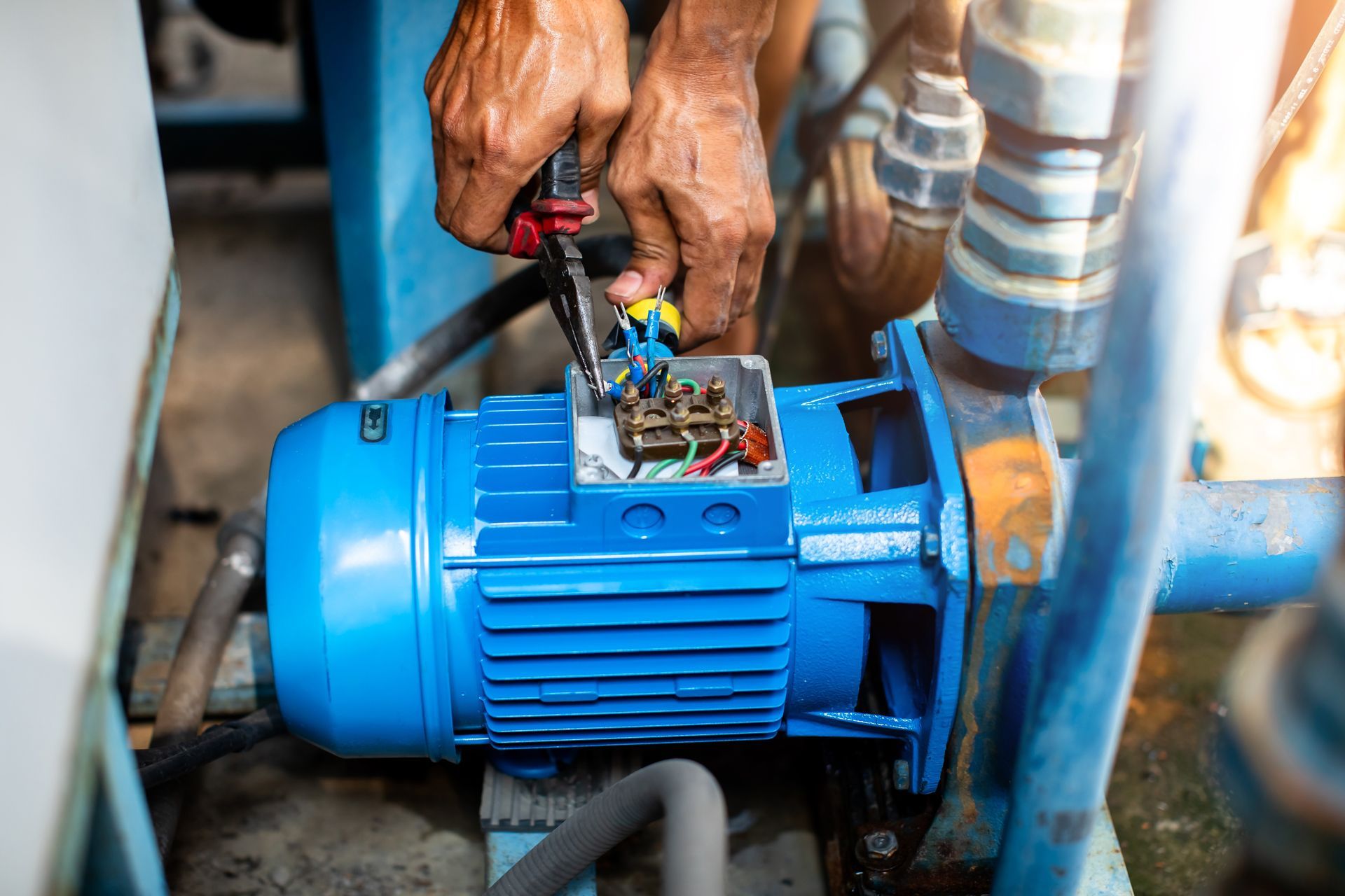 A person uses pliers to repair the electrical wiring in an open connection box on top of a blue industrial motor.