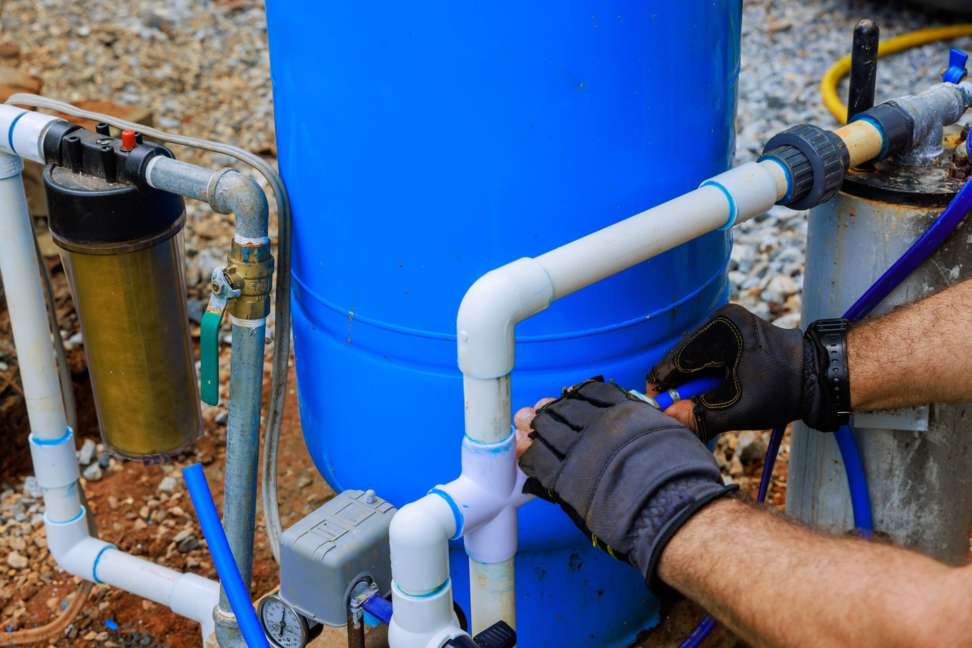 Hands in work gloves assembling white PVC piping connected to a blue water barrel and filtration system outdoors.