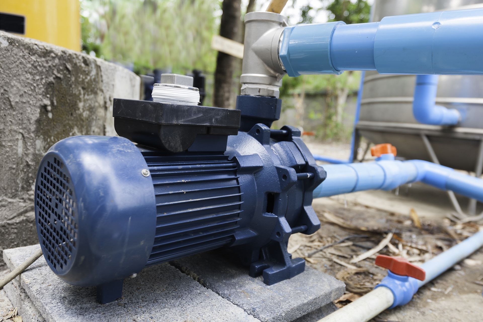 A blue electric water pump connected to blue pipes and a metal tank, installed on a concrete block base outdoors.