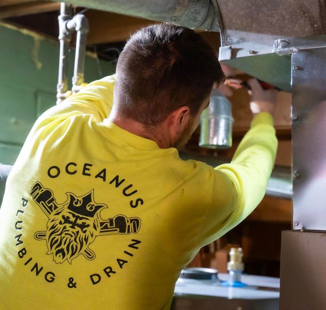 Plumber in a yellow Oceanus Plumbing & Drain shirt working under a sink in a kitchen cabinet