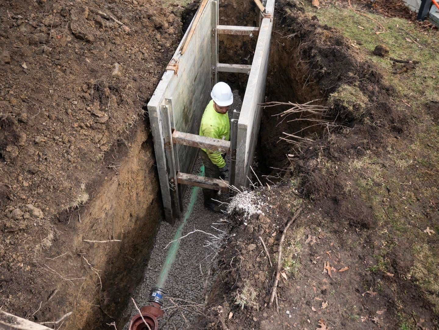 Worker in a hard hat stands in a narrow trench beside a concrete wall, spraying water on the ground.