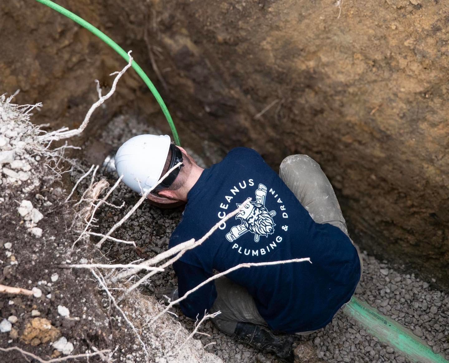 Worker in white hard hat and navy shirt crouches in a trench beside a green hose.