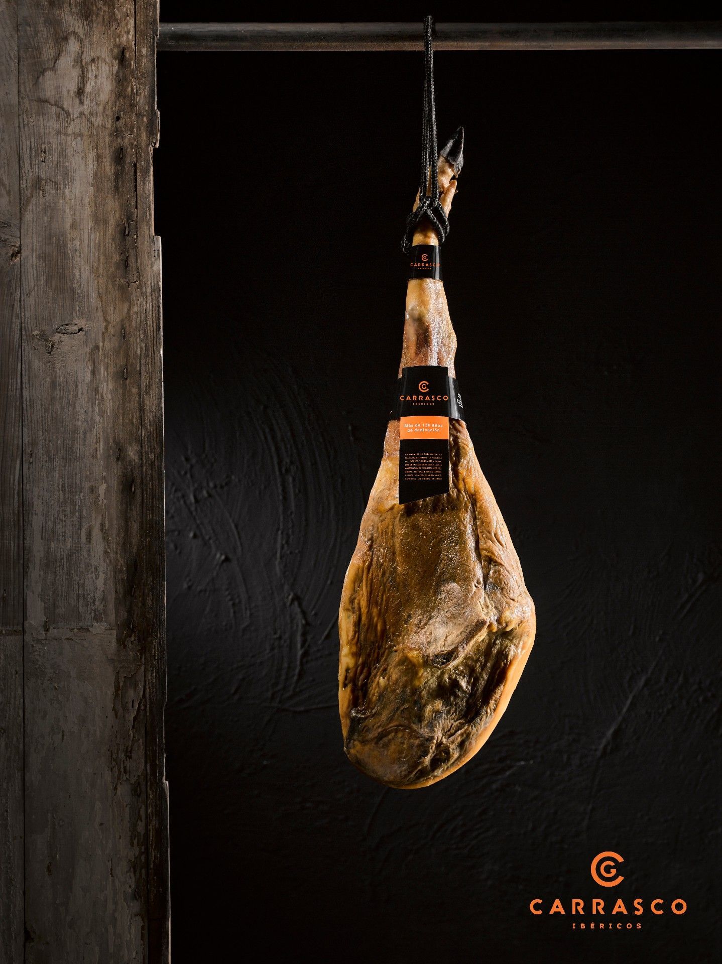 Jamón curado colgando en una habitación rústica y oscura con el logotipo de Santos Carrasco en la esquina.