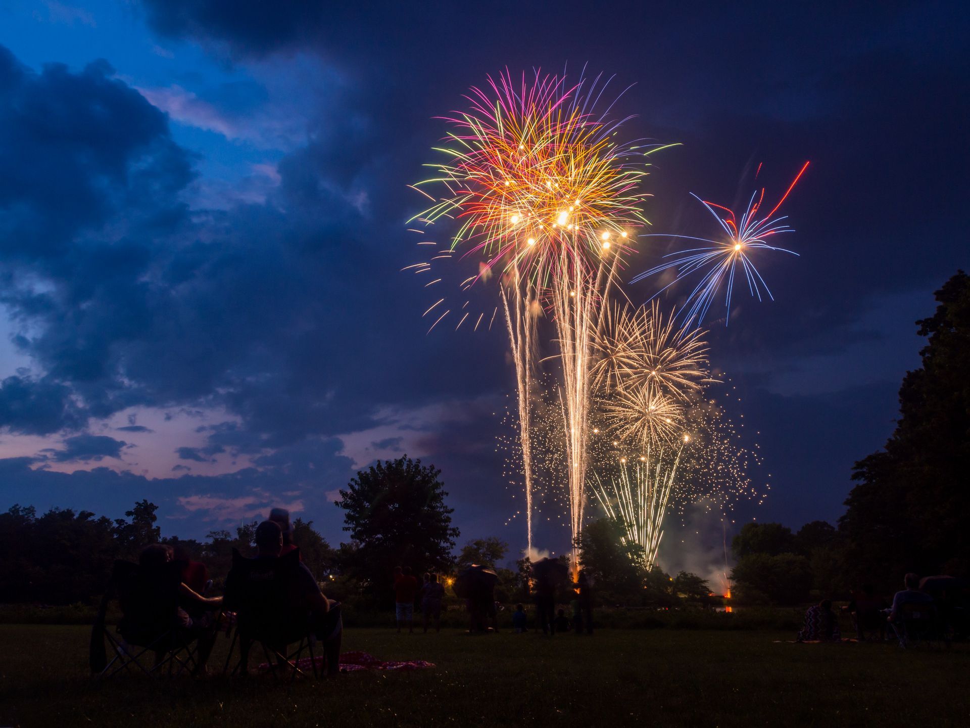 Colorful fireworks bursting over a dark field at dusk, with silhouettes of people below