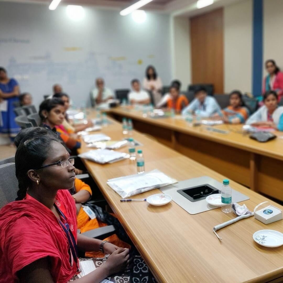 A group of people are sitting at long tables in a conference room.