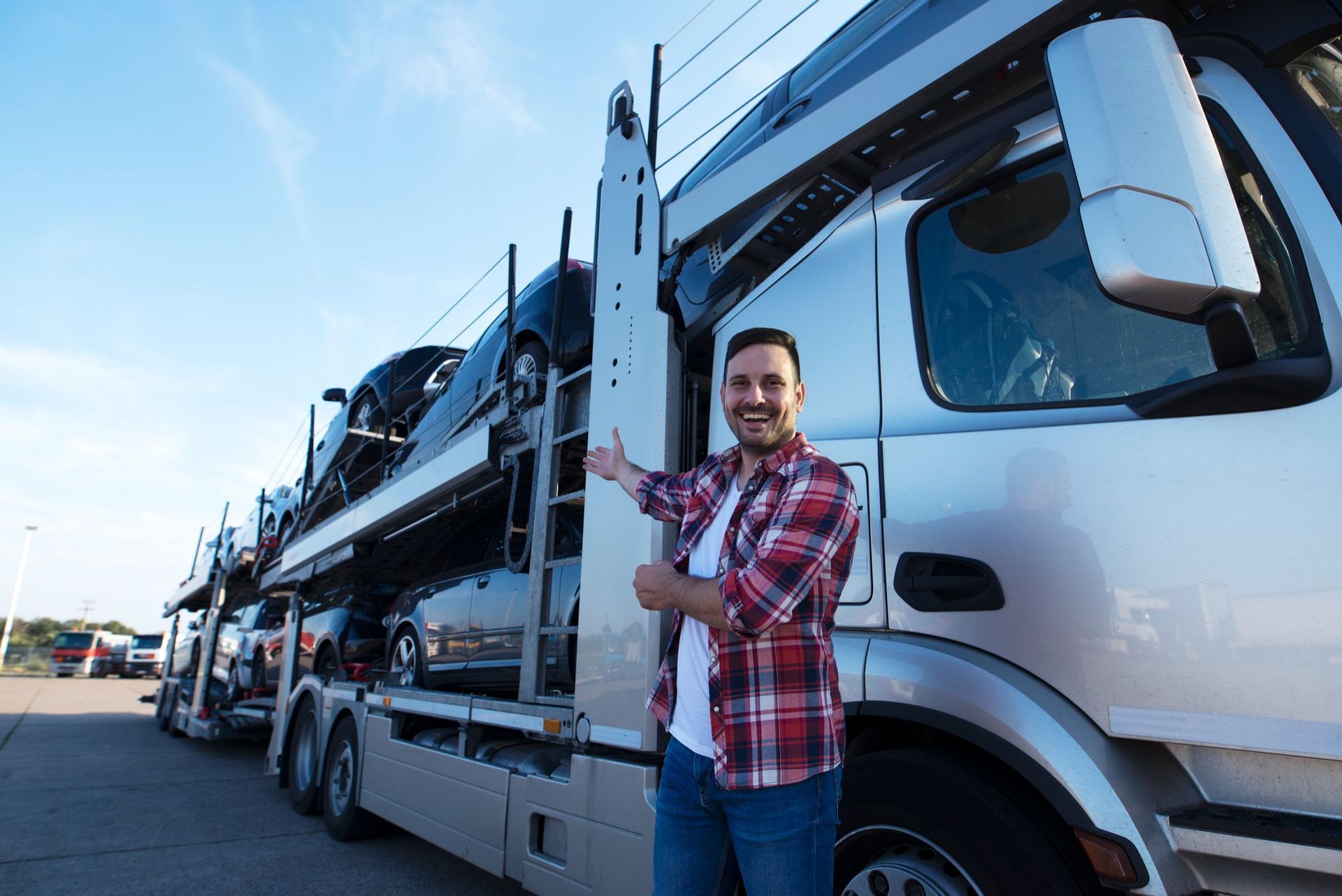 Man standing beside a car carrier truck loaded with cars on a road, smiling and pointing upward