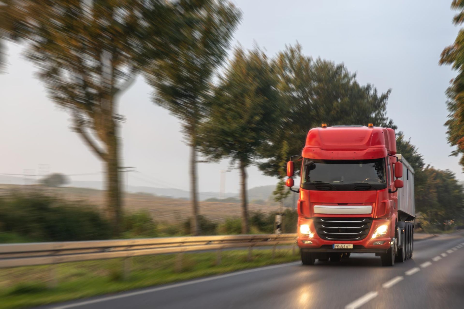 Red semi-truck driving on a highway at dusk with trees lining the road.