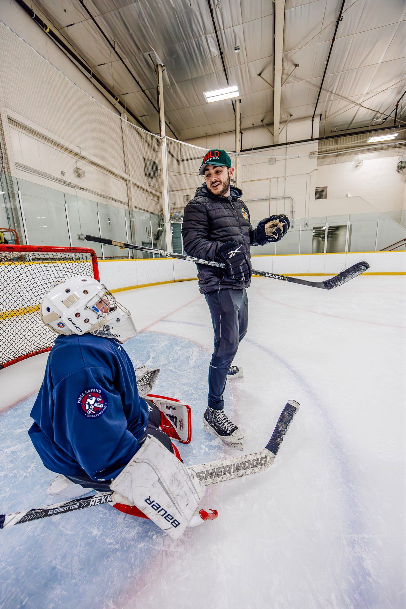 Ice hockey goalie in blue gear on the rink, with a player standing nearby holding a stick.