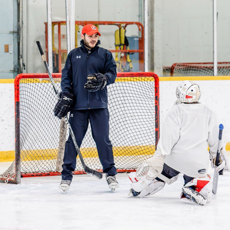 Hockey coach instructing a goalie in white gear beside the net on an indoor ice rink