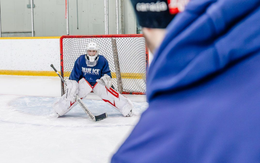 Hockey goalie in blue jersey crouches in net as a player in a blue jacket watches from the foreground