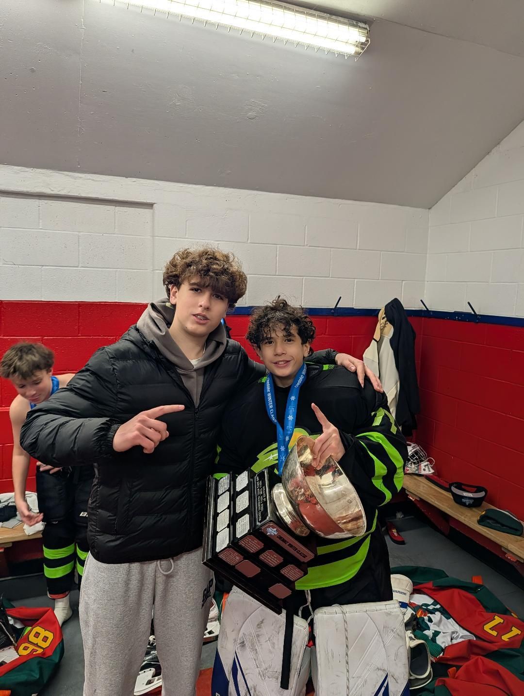 Two athletes in a locker room, one holding a medal and gift bag, with red and white walls behind them.