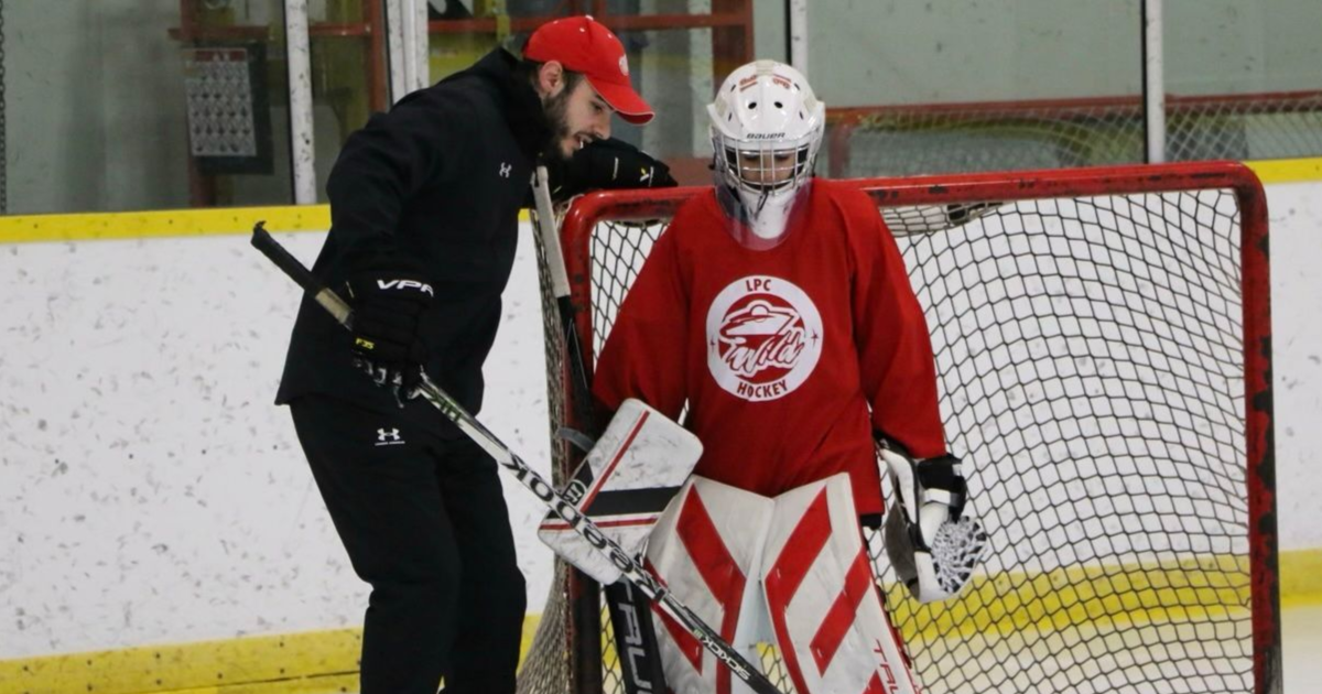 Hockey goalie in red gear on the ice beside a coach near the net