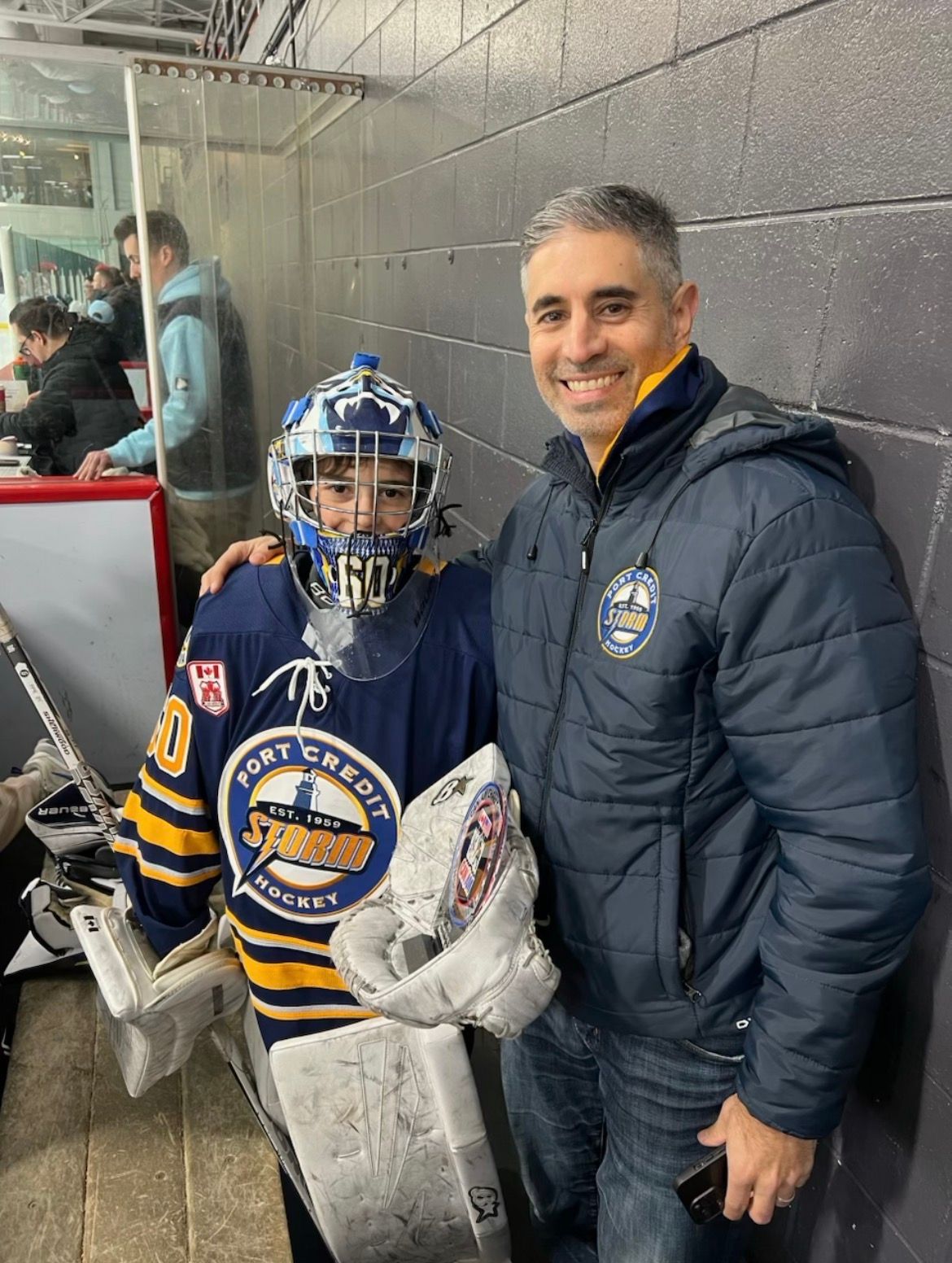 Hockey player in blue gear with goalie mask stands beside a smiling man in a rink hallway