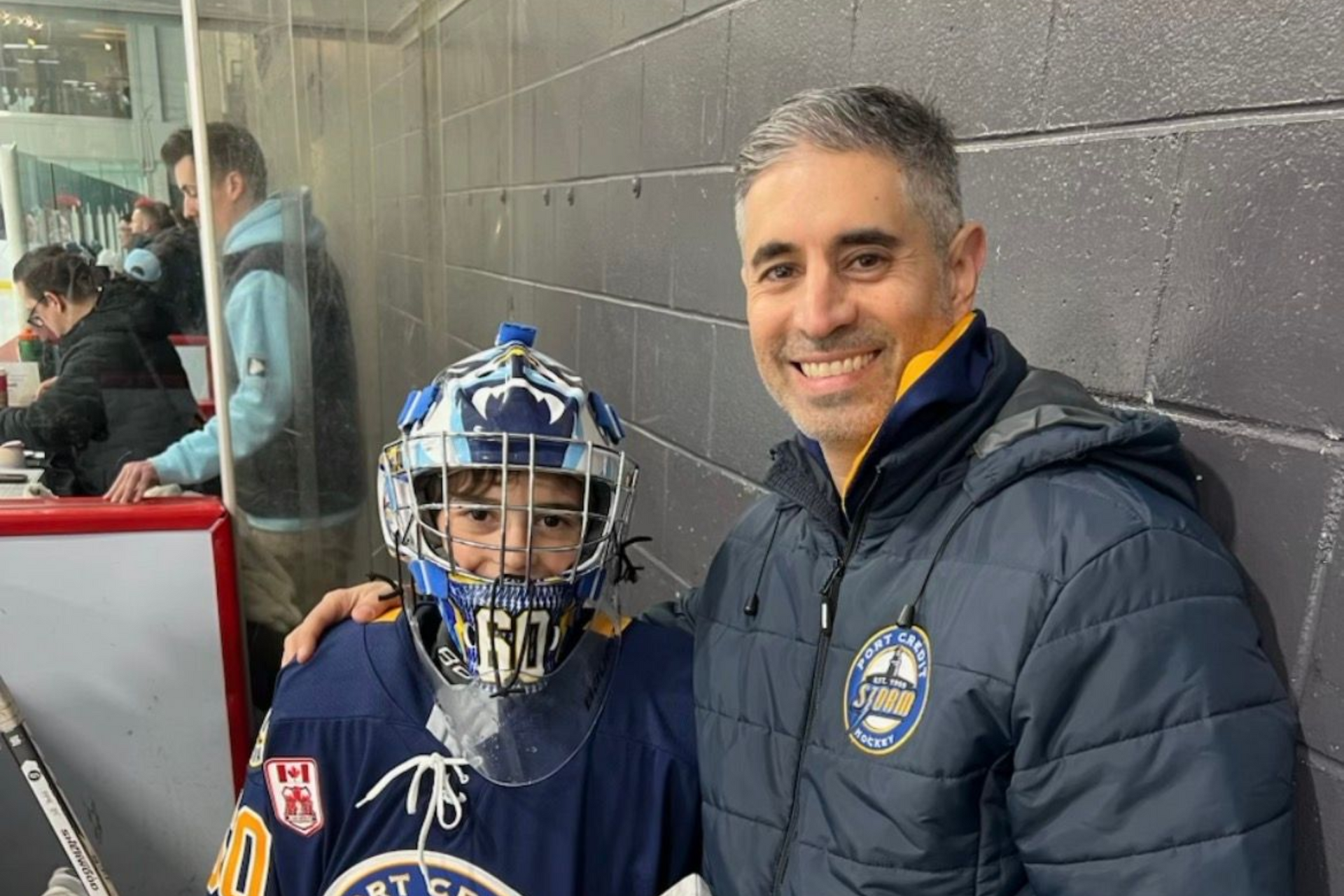 Smiling man in dark jacket poses with young hockey player in blue gear at rink side