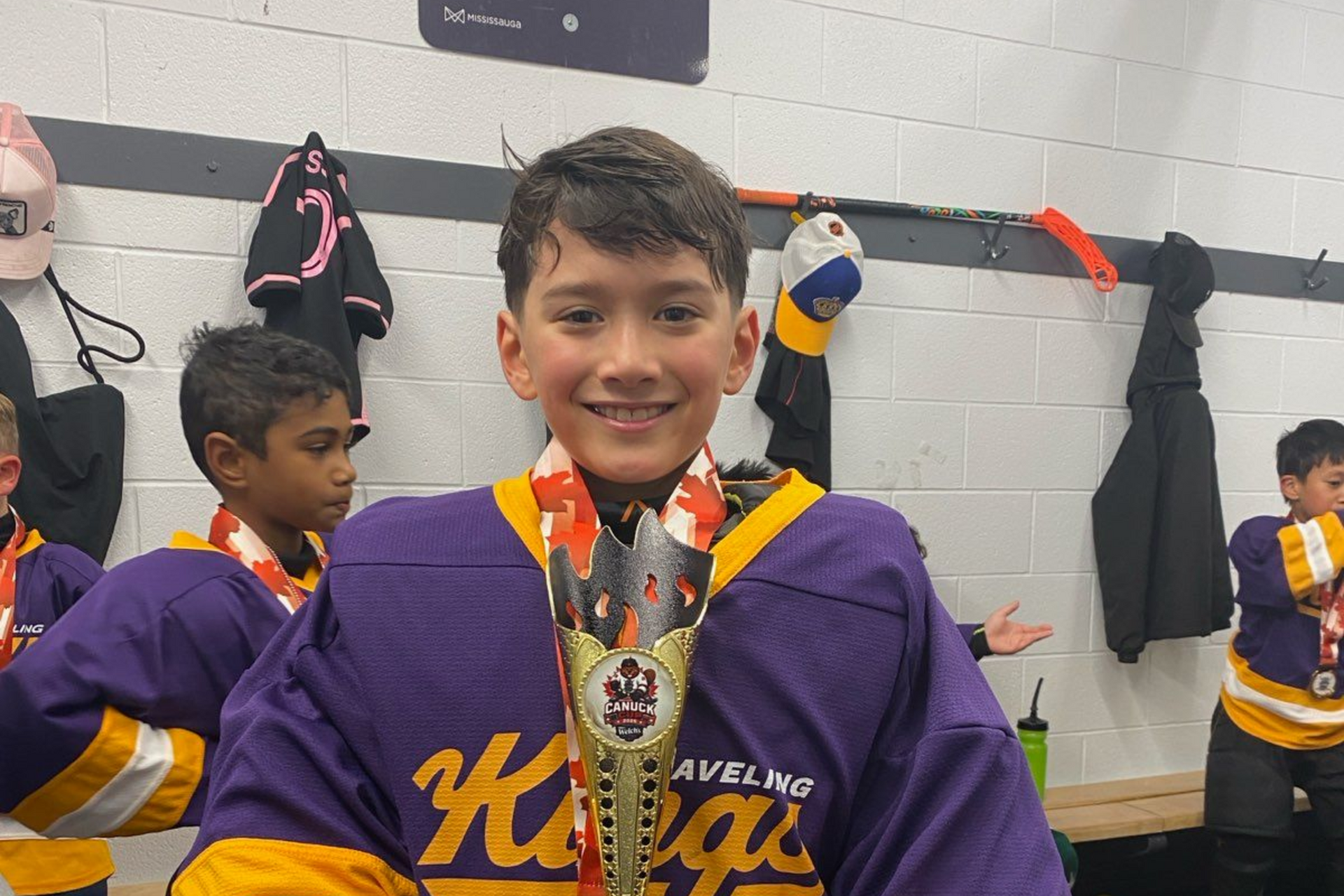 Young athlete in a purple hockey jersey smiles in a locker room with teammates and gear behind him.