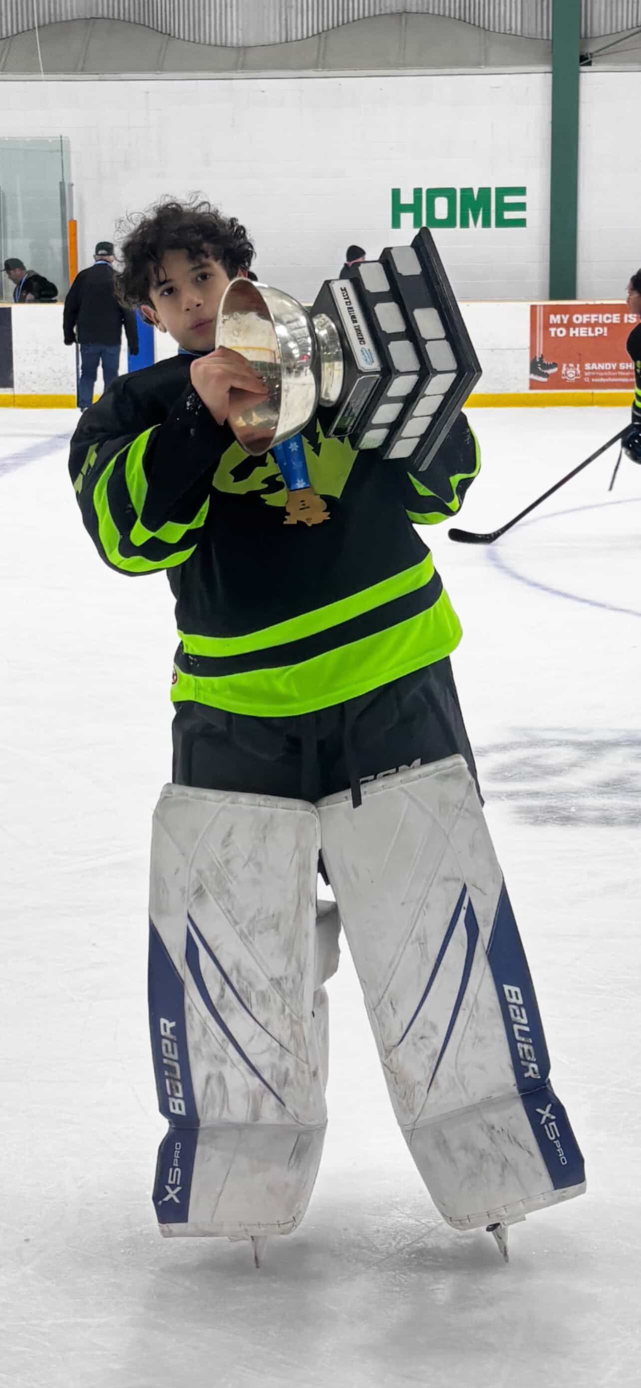 Hockey goalie in black and neon green gear holding up a trophy on an ice rink