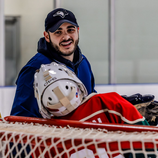 Hockey player in red gear lying near the net while a smiling man in a blue jacket stands behind him