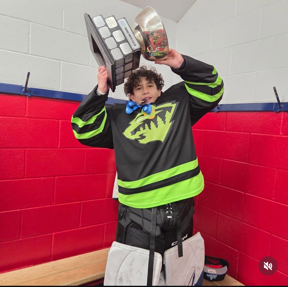 Person in a black and neon green hockey jersey holds up two trophies in a locker room.