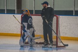 Hockey goalie in blue gear stands beside a coach on the ice in front of the net.