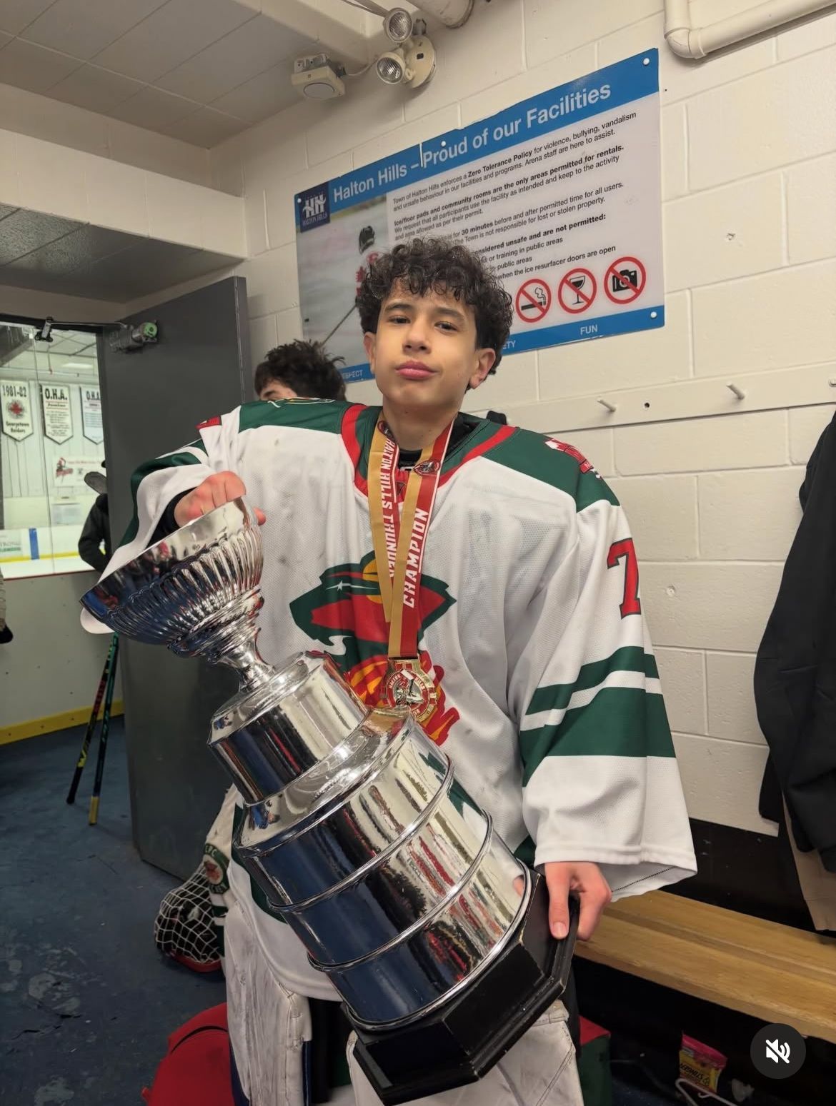 Person in a white hockey jersey holds a large silver trophy in a locker room