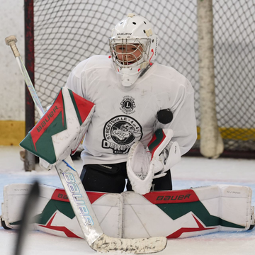 Hockey goalie in white gear and red-green pads kneels in front of the net on the ice.